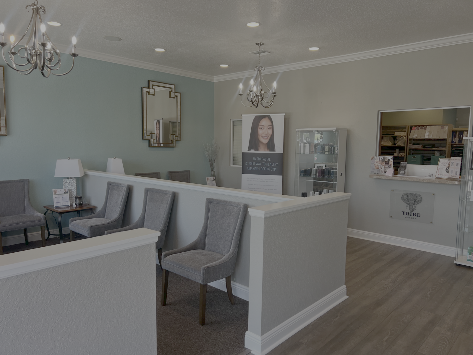 The reception and waiting area at Tribe Med Spa Lutz — grey upholstered chairs beside a half-height partition wall, a lit consultation desk, and pendant chandeliers overhead